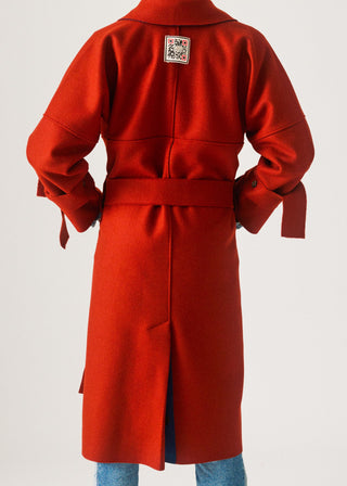 A bright red wool-blend coat shown from the back, featuring a self-tie belt, long sleeves with buttoned cuffs, and a small decorative patch on the upper back. The coat is modeled on a person wearing blue jeans against a plain light gray studio background.