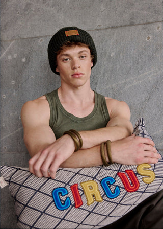 Person wearing a green tank top and black beanie sitting on a cushion with 'CIRCUS' text against a gray wall.