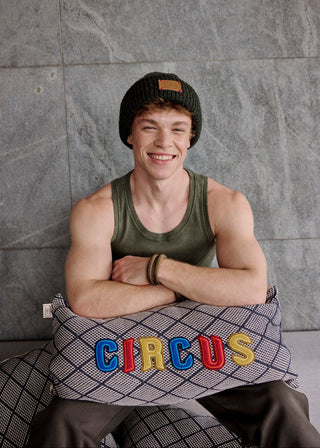 Person wearing a tank top and beanie sitting on a pillow with 'CIRCUS' letters against a gray wall.