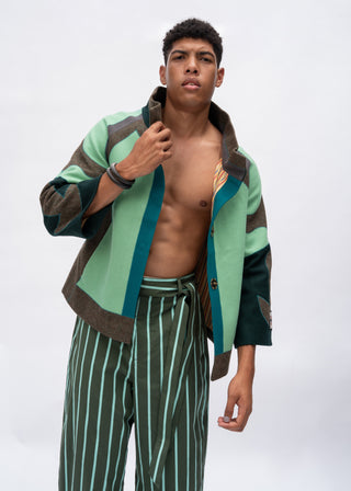 A male model wearing a color-blocked knit cardigan in sage green, brown, and teal, paired with green and white vertically striped trousers, against a plain light gray studio background.