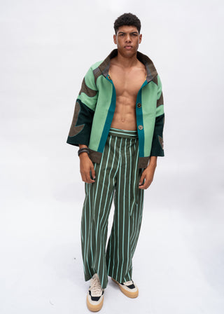 A male model stands against a white studio background wearing a cropped, open-front jacket with sage green, dark green, and brown color-blocked panels, matching green-and-white vertically striped wide-leg pants, and beige-and-black sneakers.
