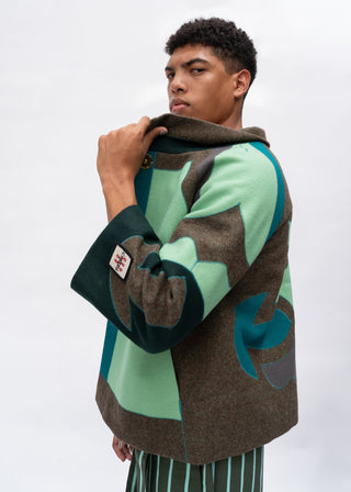 A model wearing a color-blocked knit cardigan with sage green, brown, and dark teal panels, paired with green and white striped trousers, against a light gray studio background.