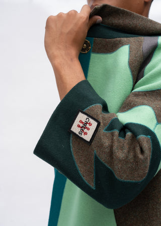 Close-up of a person wearing a patterned wool coat with a visible Cactus Jack patch on the cuff, showing the hand adjusting the collar against a plain light background.