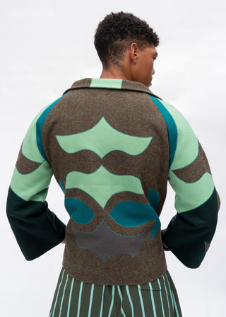 A model shown from the back wearing a brown wool-blend cardigan with large abstract green and teal patches, paired with green and white striped trousers, against a light gray studio background.