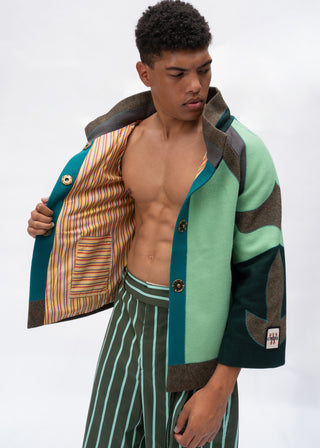 A male model wearing a sage green and dark teal patchwork wool-blend jacket with brown suede-like trim, shown open to reveal a striped lining. He is also wearing green and white vertically striped trousers, photographed against a clean white studio background.