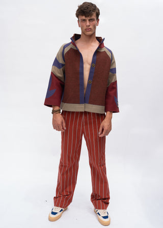 A male model stands against a white studio background wearing a matching two-piece outfit consisting of a color-blocked zip-up hoodie and red pinstriped trousers.