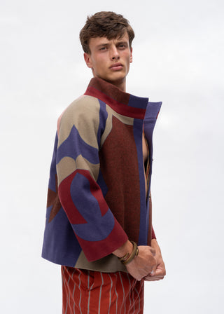 A male model wearing a colorful geometric patterned wool-blend jacket with red, blue, and beige shapes, paired with red and white striped trousers, against a plain light gray background.