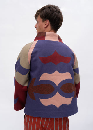 A man shown from the back wearing a colorful patchwork wool jacket with abstract geometric shapes in purple, beige, red, and brown, paired with red and white striped trousers, against a plain light gray background.