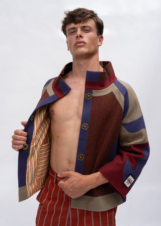 A male model wearing a color-blocked knit cardigan with red, blue, and beige panels, worn open to show the striped lining, paired with red and white striped trousers against a light gray background.